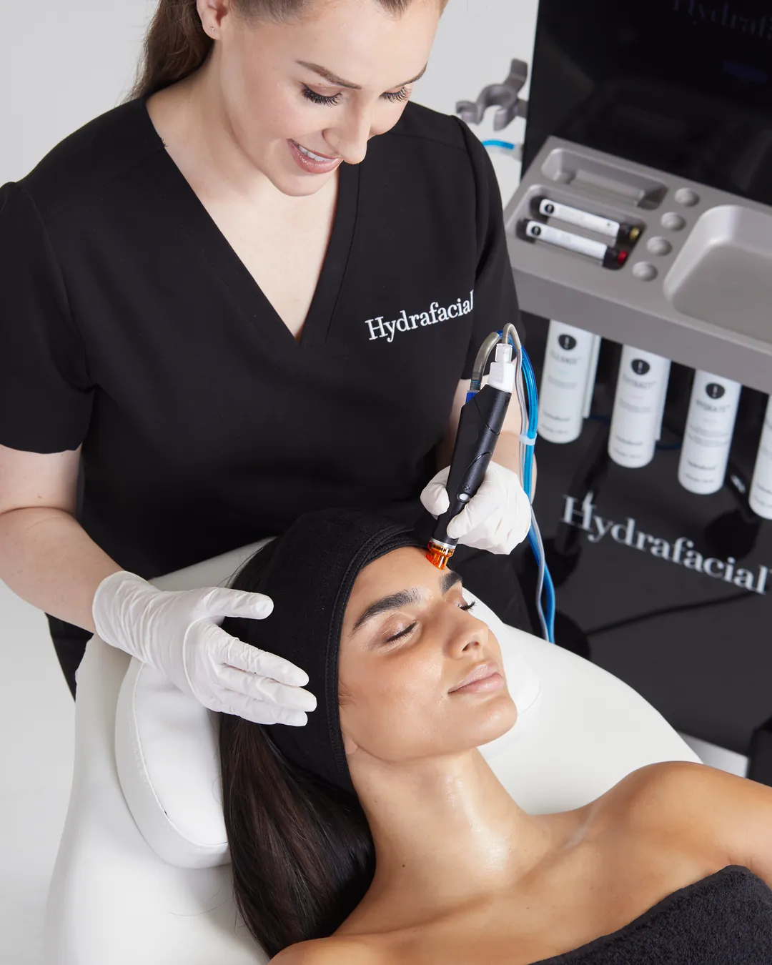 Specialistă zâmbitoare care lucrează cu piesa de mână pe fruntea unei paciente relaxate, cu părul prins sub o banderolă, lângă aparatul Hydrafacial Syndeo
