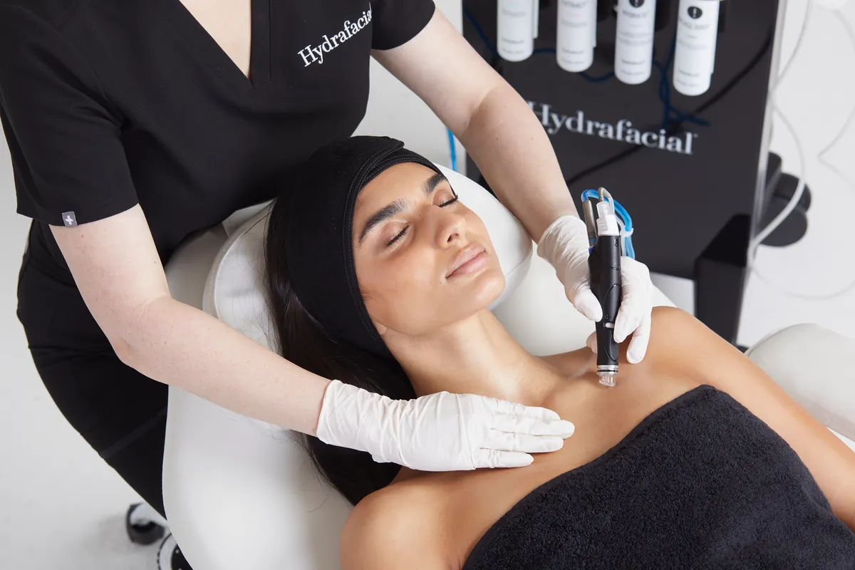 Specialistă cu mănuși care trece piesa de mână Hydrafacial pe decolteul unei paciente întinse pe pat, lângă aparat