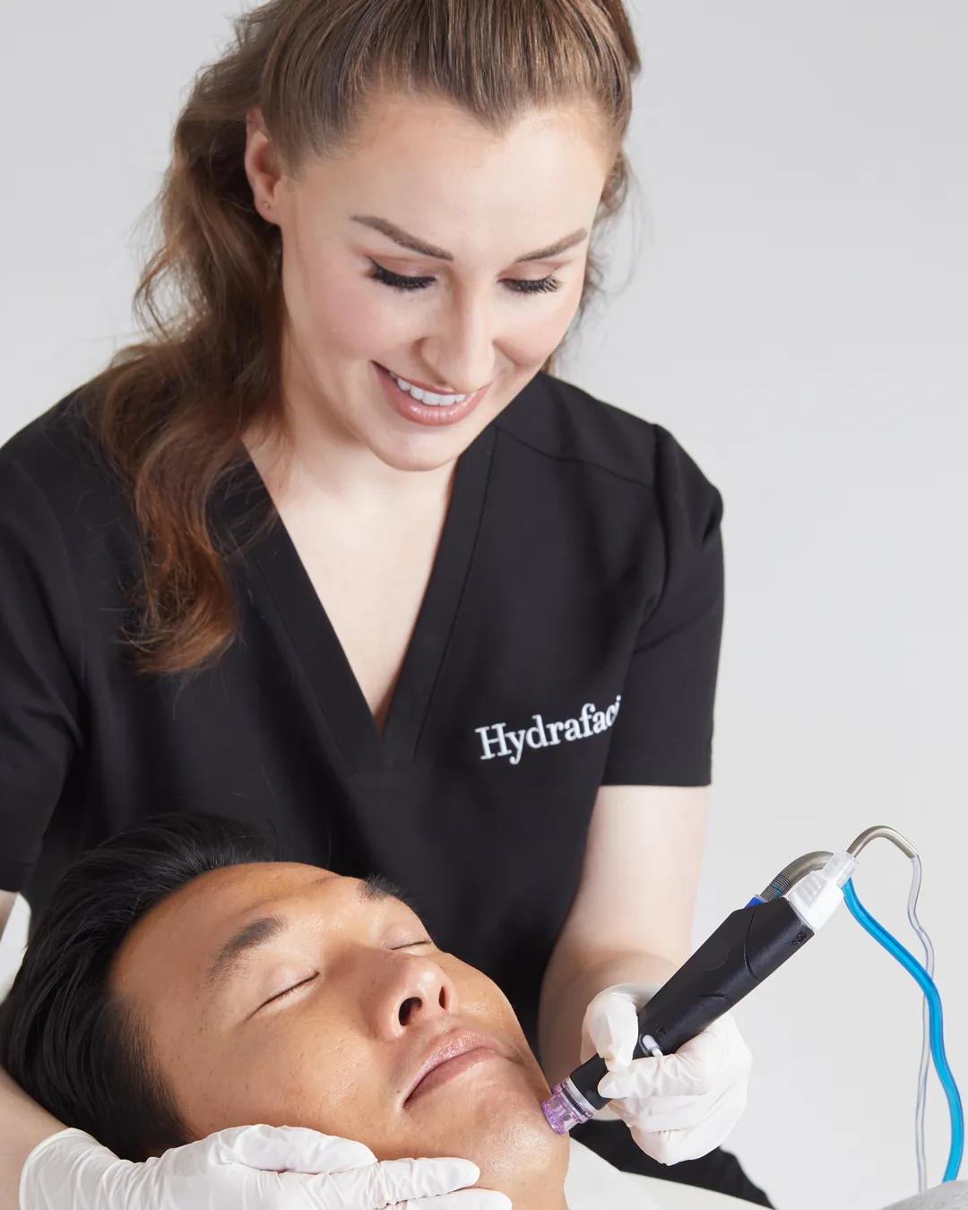 Bărbat întins pe patul de tratament, cu ochii închiși, în timp ce o specialistă zâmbitoare îi trece vârful piesei de mână Hydrafacial pe bărbie