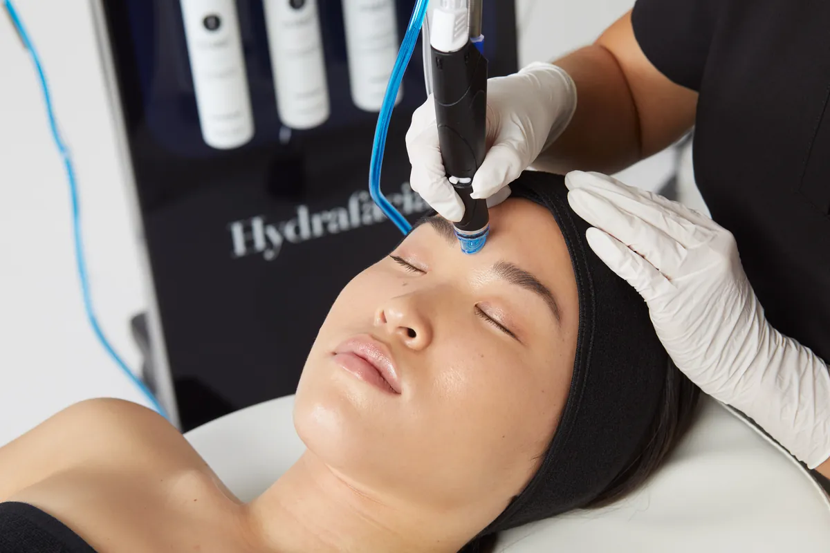 Vârful albastru, spiralat, al piesei de mână Hydrafacial pe fruntea unei femei cu ochii închiși; în fundal, aparatul și flacoanele de soluții